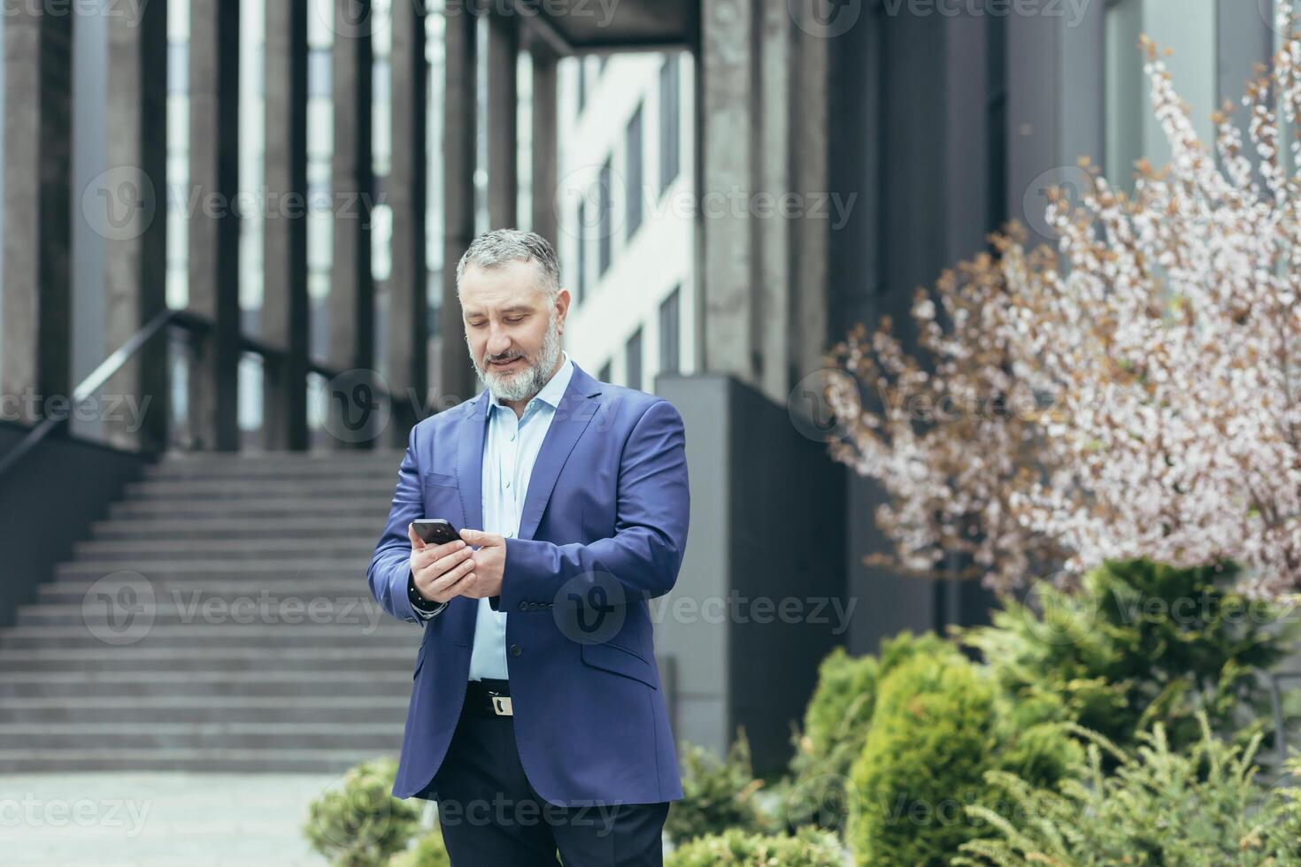 Senior grisalho homem, uma homem de negocios dentro uma terno, carrinhos perto a moderno escritório Centro e chamadas uma Táxi a partir de a telefone, digitando a número, espera, é dentro uma pressa para encontrar. foto