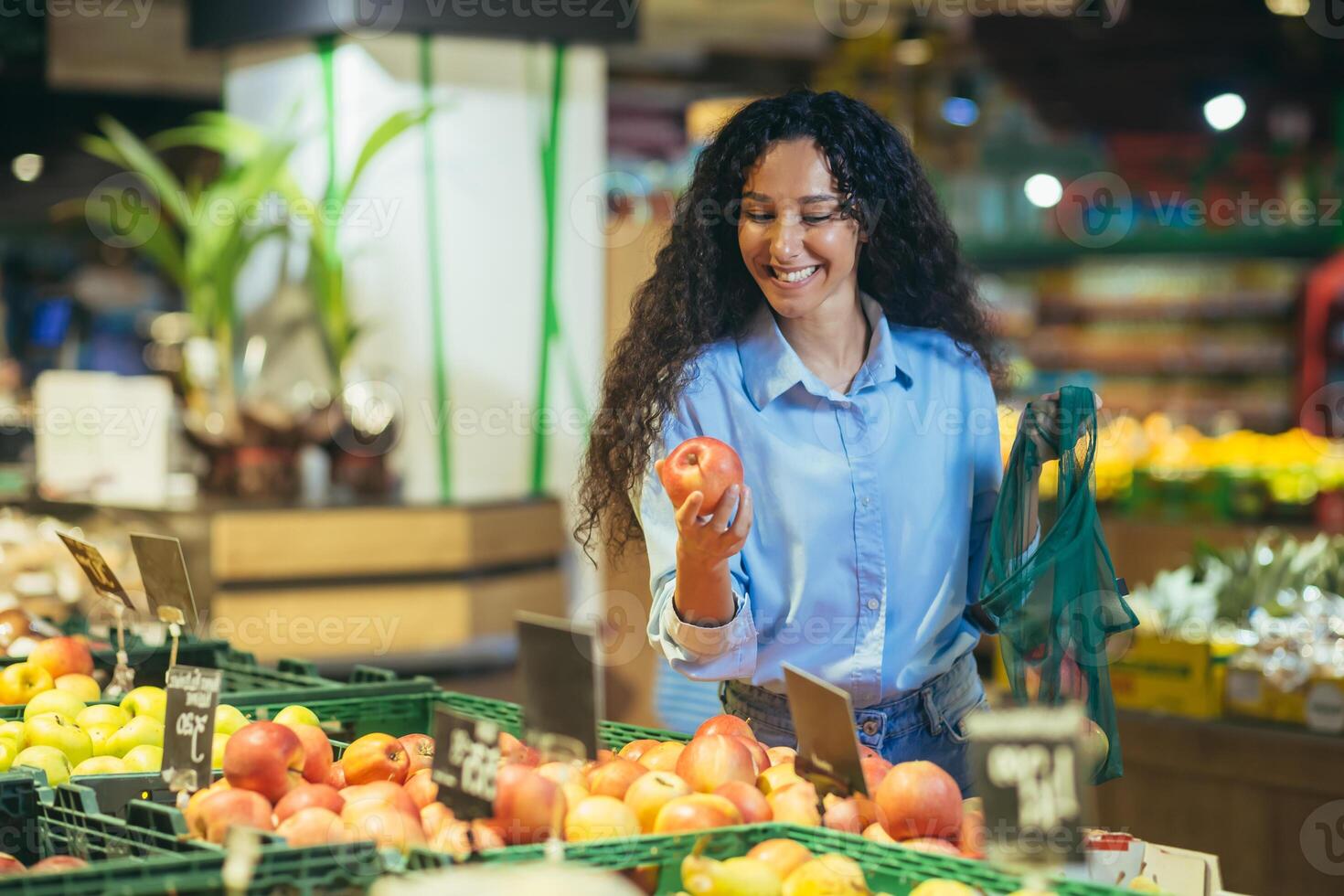 uma jovem hispânico mulher é compras dentro uma supermercado. picaretas e detém a maçã, coloca dentro a Bolsa ecologica foto