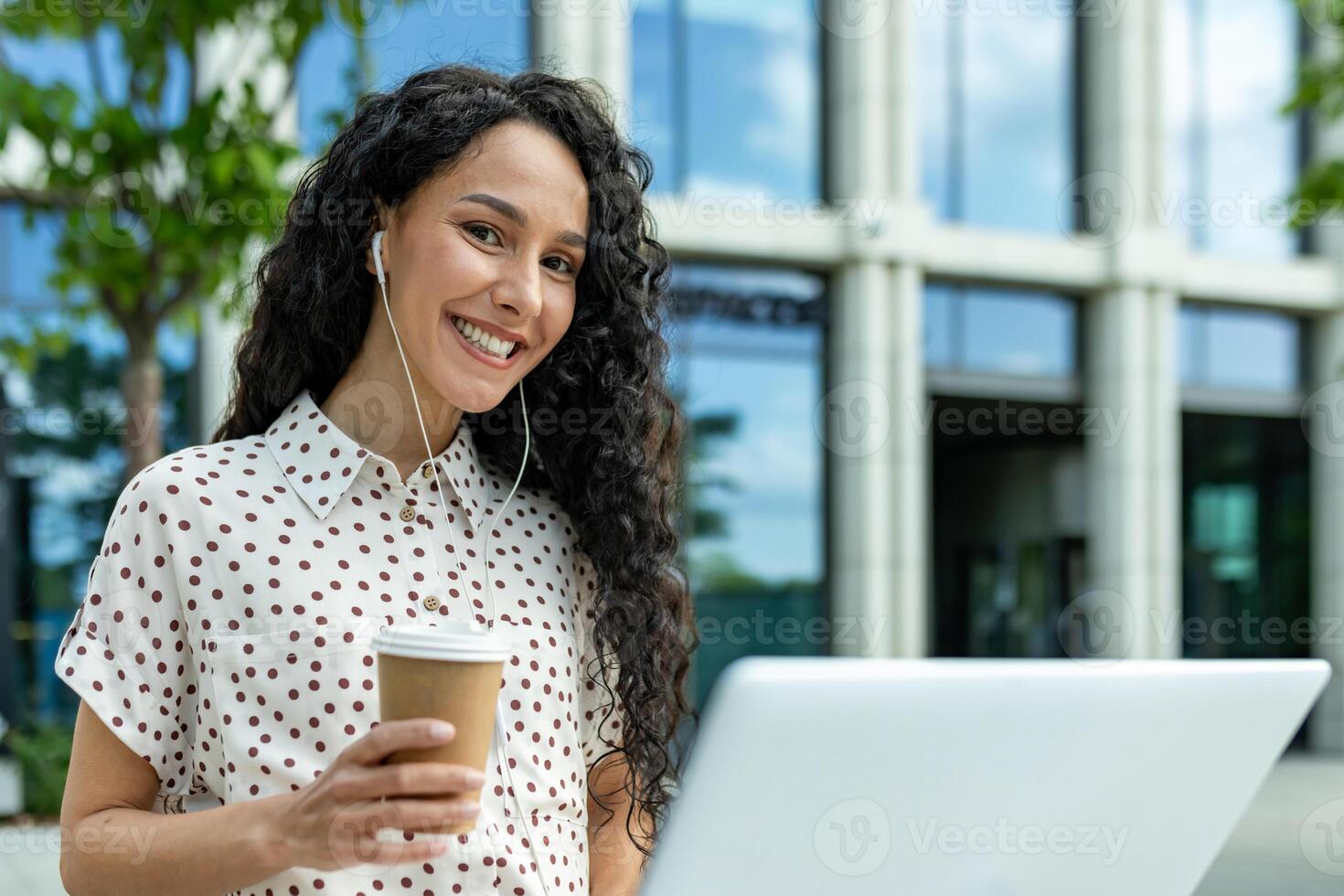 sorridente jovem mulher com fones de ouvido e café levando uma pausa a partir de trabalhos em dela computador portátil lado de fora uma moderno escritório prédio. foto