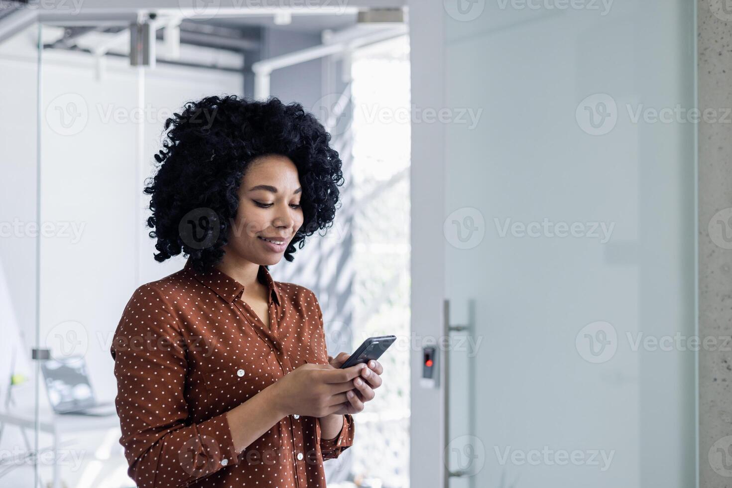 uma profissional africano americano mulher dentro uma à moda polca ponto blusa usa dela Smartphone dentro uma brilhante, moderno escritório ambiente, olhando conteúdo e focado. foto