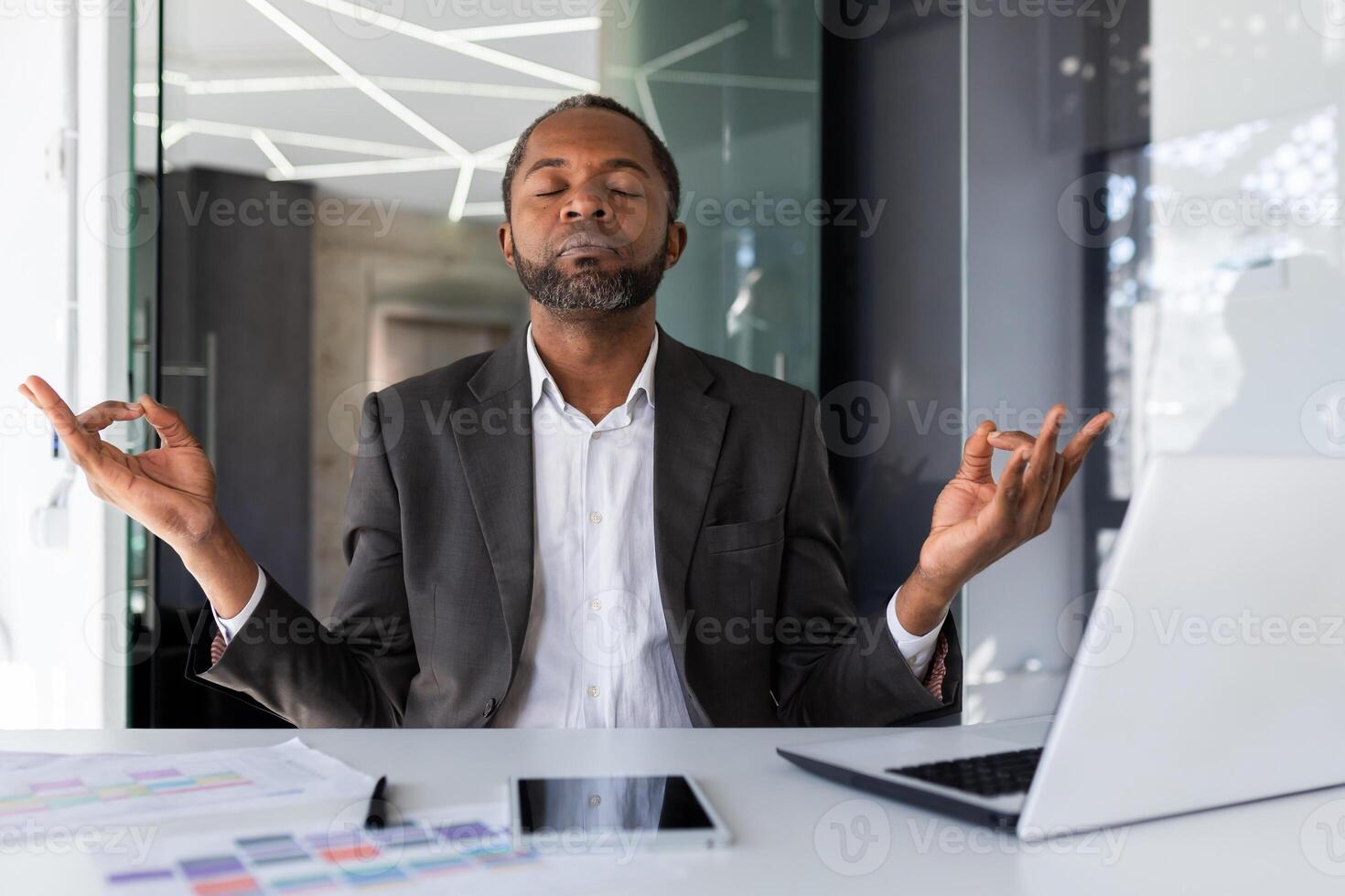maduro com experiência africano americano patrão meditando dentro escritório local de trabalho sentado às mesa, homem com fechadas olhos sentado visualizando futuro vitórias e conquistas, homem de negocios investidor com computador portátil. foto