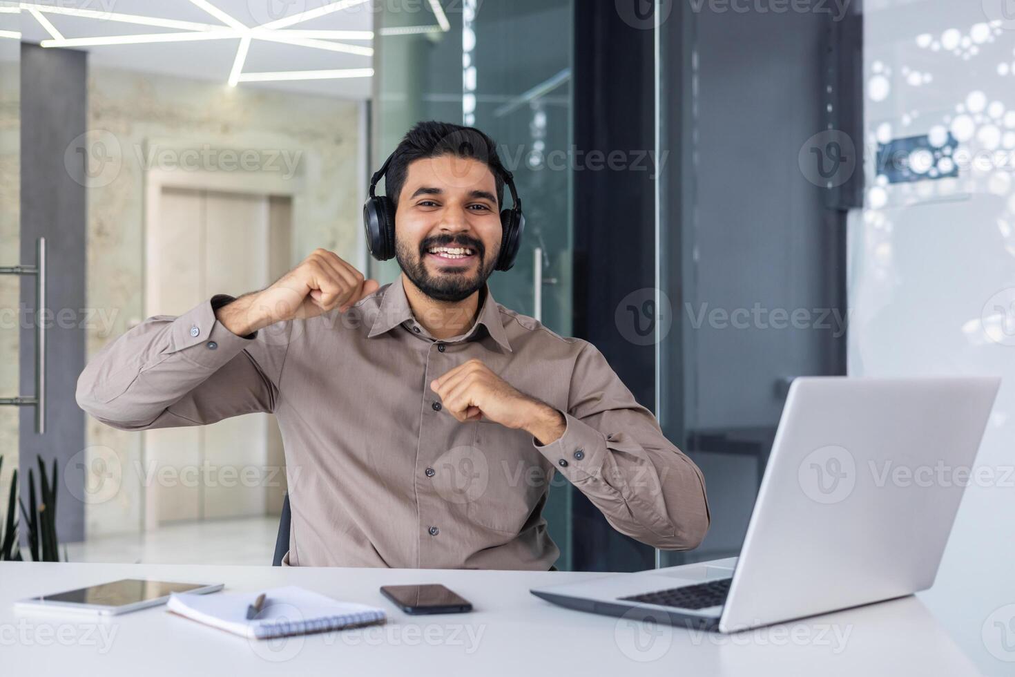 retrato do uma feliz jovem indiano homem sentado às uma escrivaninha dentro a escritório, trabalhando em uma computador portátil, olhando sorridente às a Câmera e dançando dentro alegria do sucesso. foto