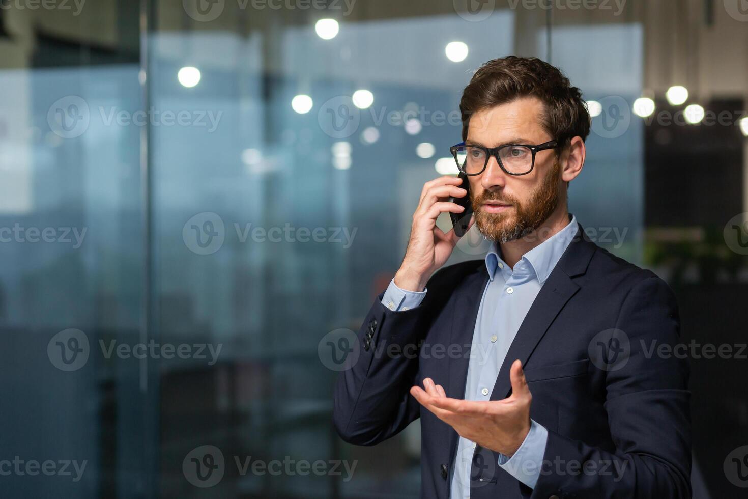 sério e bem sucedido maduro homem de negocios dentro escritório perto janela falando em telefone, homem dentro o negócio terno e barba de pé. foto