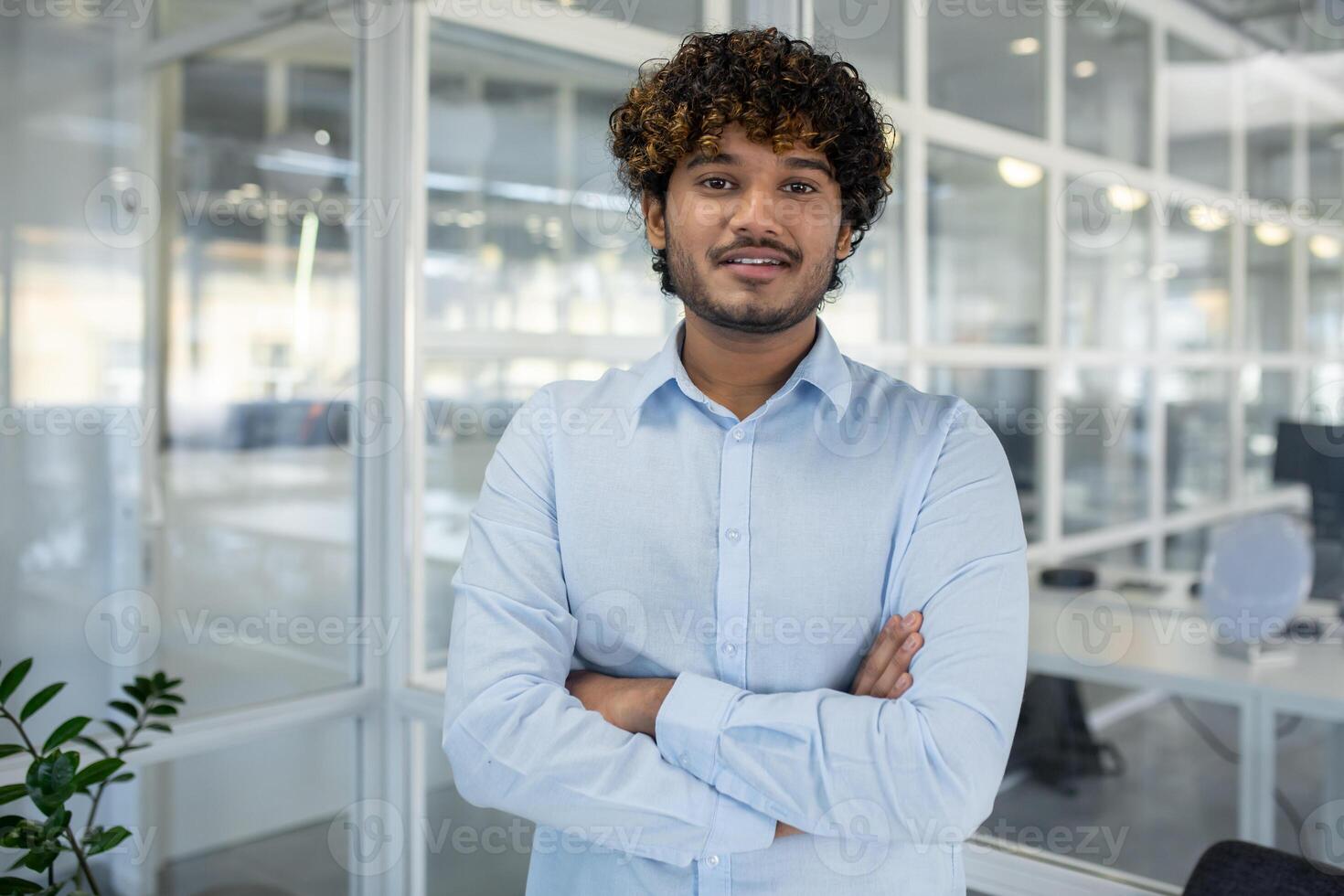 uma retrato do uma confiante jovem homem de negocios com encaracolado cabelo, em pé braços cruzado dentro uma cheio de luz, contemporâneo escritório ambiente. foto