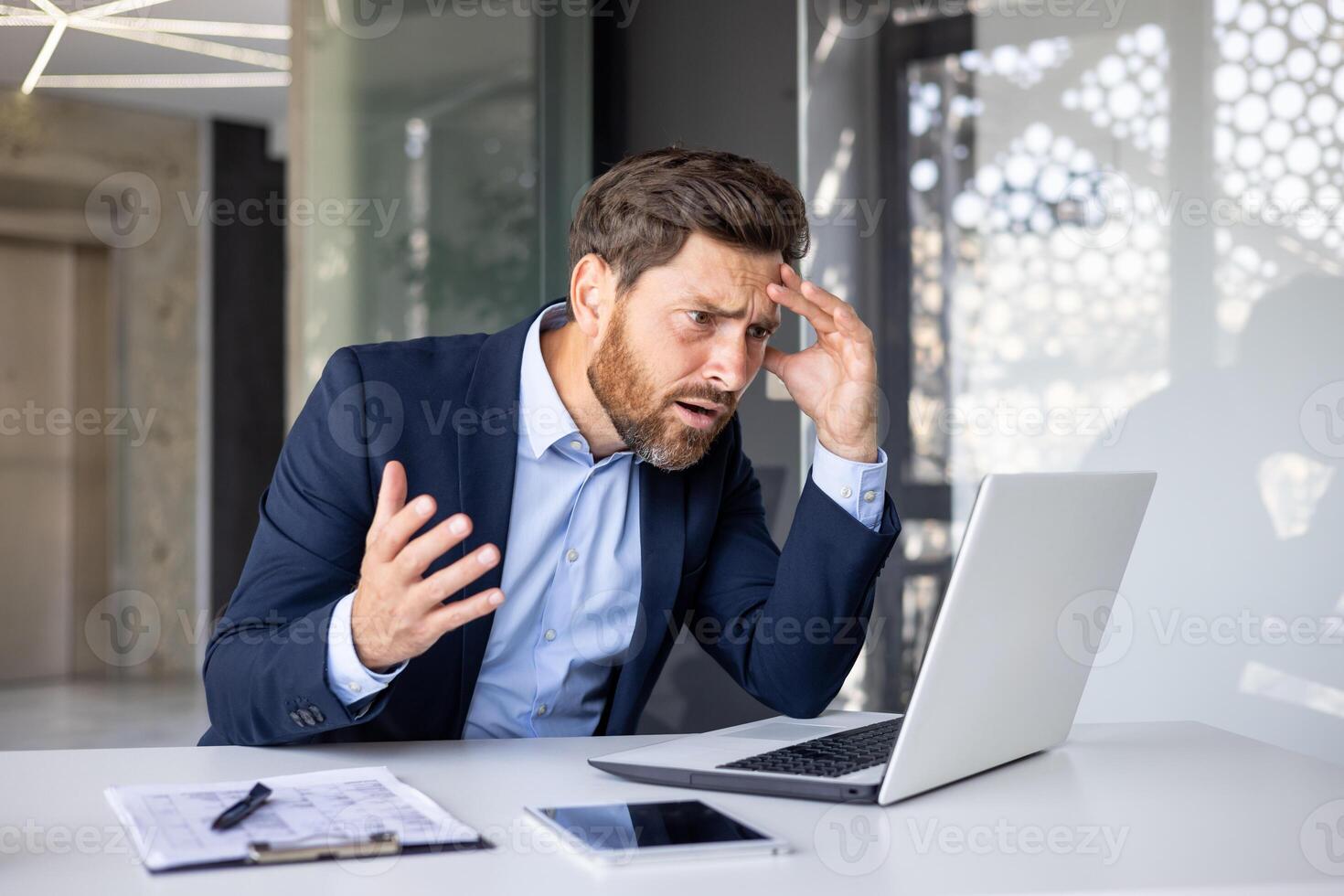 chocado e surpreso jovem homem de negocios homem sentado às escrivaninha dentro escritório e olhando a sério às computador portátil monitor segurando cabeça. foto