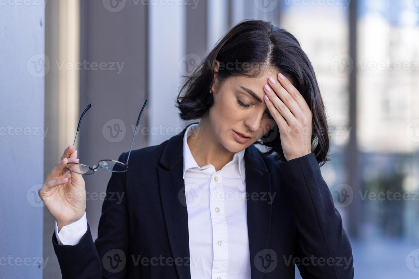 jovem mulher cansado com dor de cabeça, empresária lado de fora escritório construção esfrega olhos, tonto, sobrecarregado trabalhador ao ar livre dentro o negócio terno fechar acima. foto