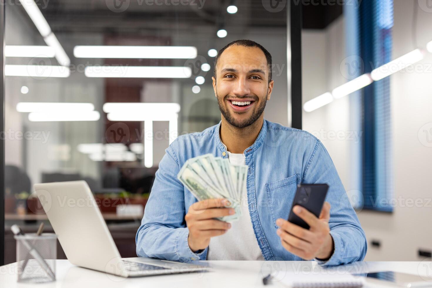 retrato do uma feliz jovem hispânico homem sentado dentro a escritório às a mesa, segurando dinheiro dinheiro e uma telefone, sorridente às a Câmera. foto