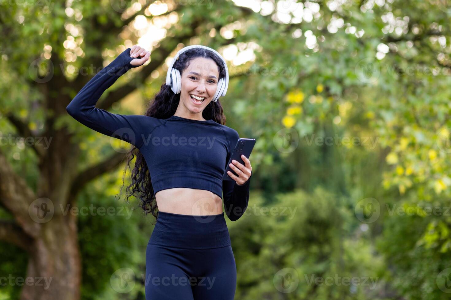 feliz mulher dentro Preto roupa ativa e branco fones de ouvido levantando mão com cerrado punho enquanto usando Smartphone. desportivo fêmea com encaracolado cabelo alcançando objetivo do corrida maratona em público parque fundo. foto