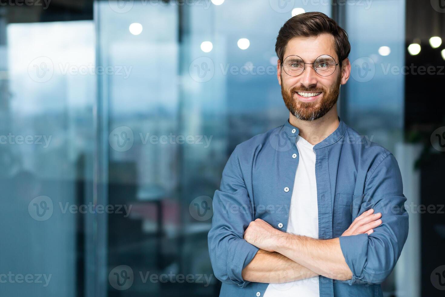 retrato do bem sucedido homem de negocios dentro moderno escritório, maduro homem com barba e óculos sorridente e olhando às Câmera, patrão dentro casual roupas camisa perto janela com cruzado braços. foto