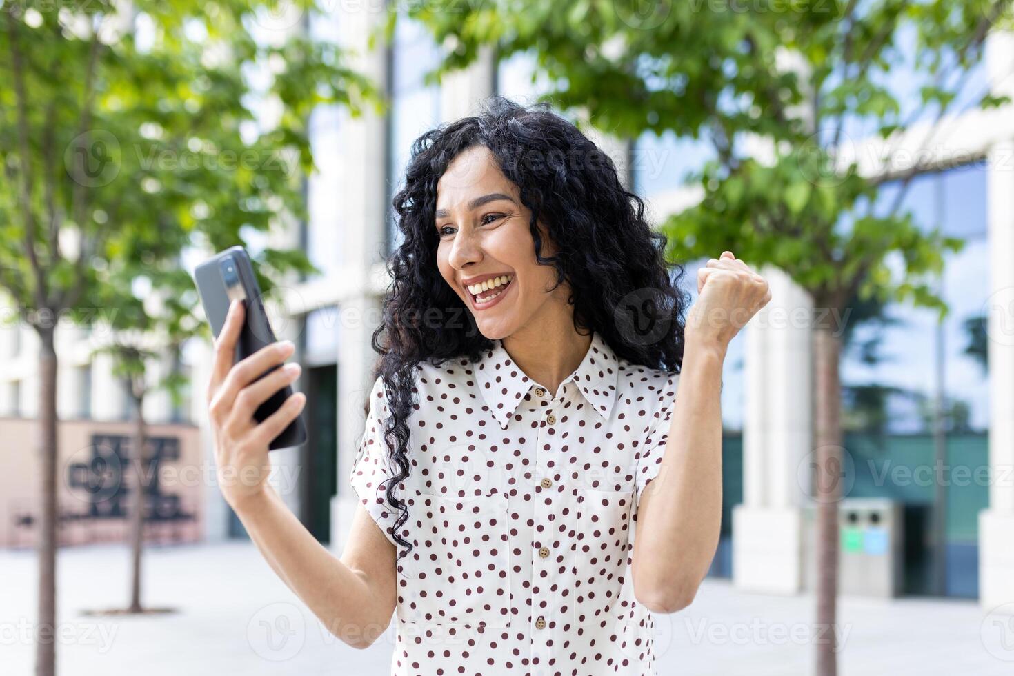 jovem alegre mulher vencedora recebido conectados notificação em telefone, hispânico mulher com encaracolado cabelo a comemorar sucesso e triunfo caminhando dentro cidade perto escritório construção fora. foto