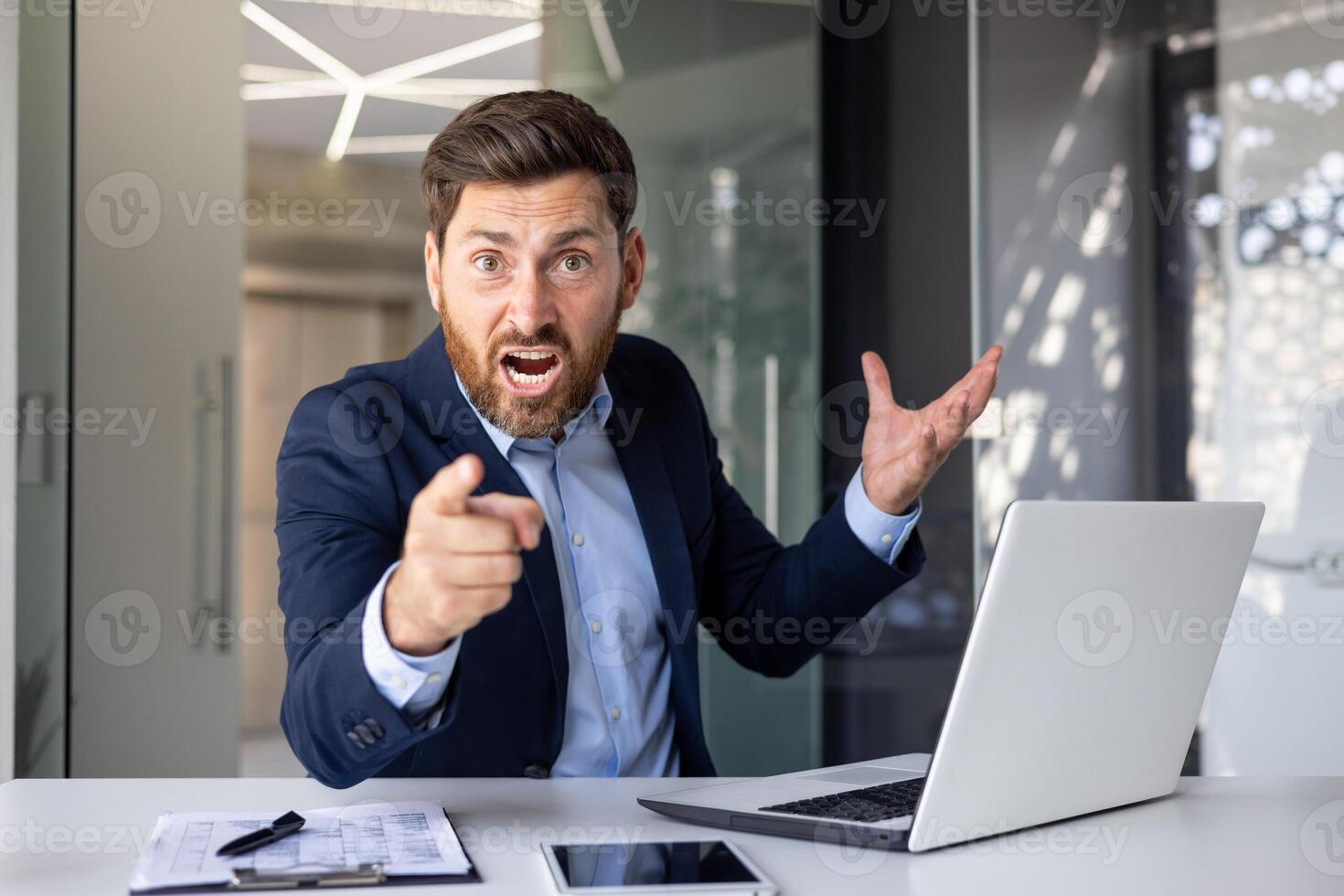 Bravo jovem homem homem de negocios sentado dentro a escritório às a mesa e apontando dele dedo às a Câmera, obtendo usava para a problema e erro, gritando descontente. foto