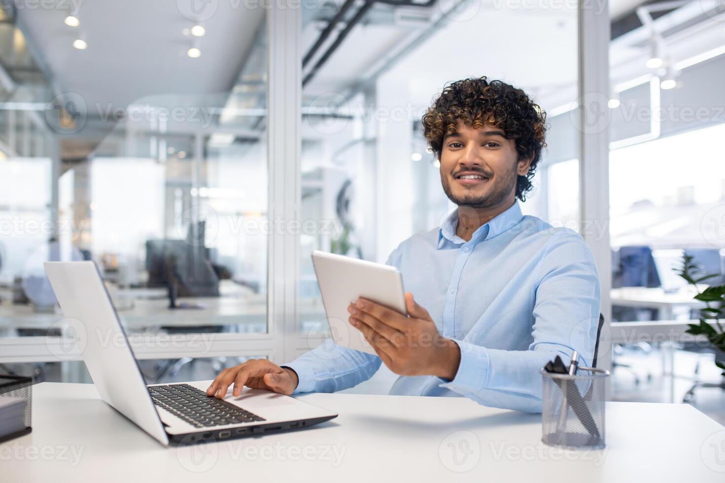 confiante jovem homem de negocios com encaracolado cabelo sorridente enquanto usando uma tábua e computador portátil às dele branco escrivaninha dentro uma brilhante contemporâneo escritório espaço. foto