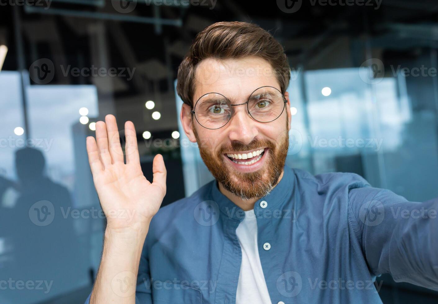 maduro homem dentro escritório vestindo camisa e óculos sorridente e olhando às Smartphone Câmera, homem de negocios levando selfie e falando em ligar usando Smartphone, programador acenando mão cumprimento gesto. foto