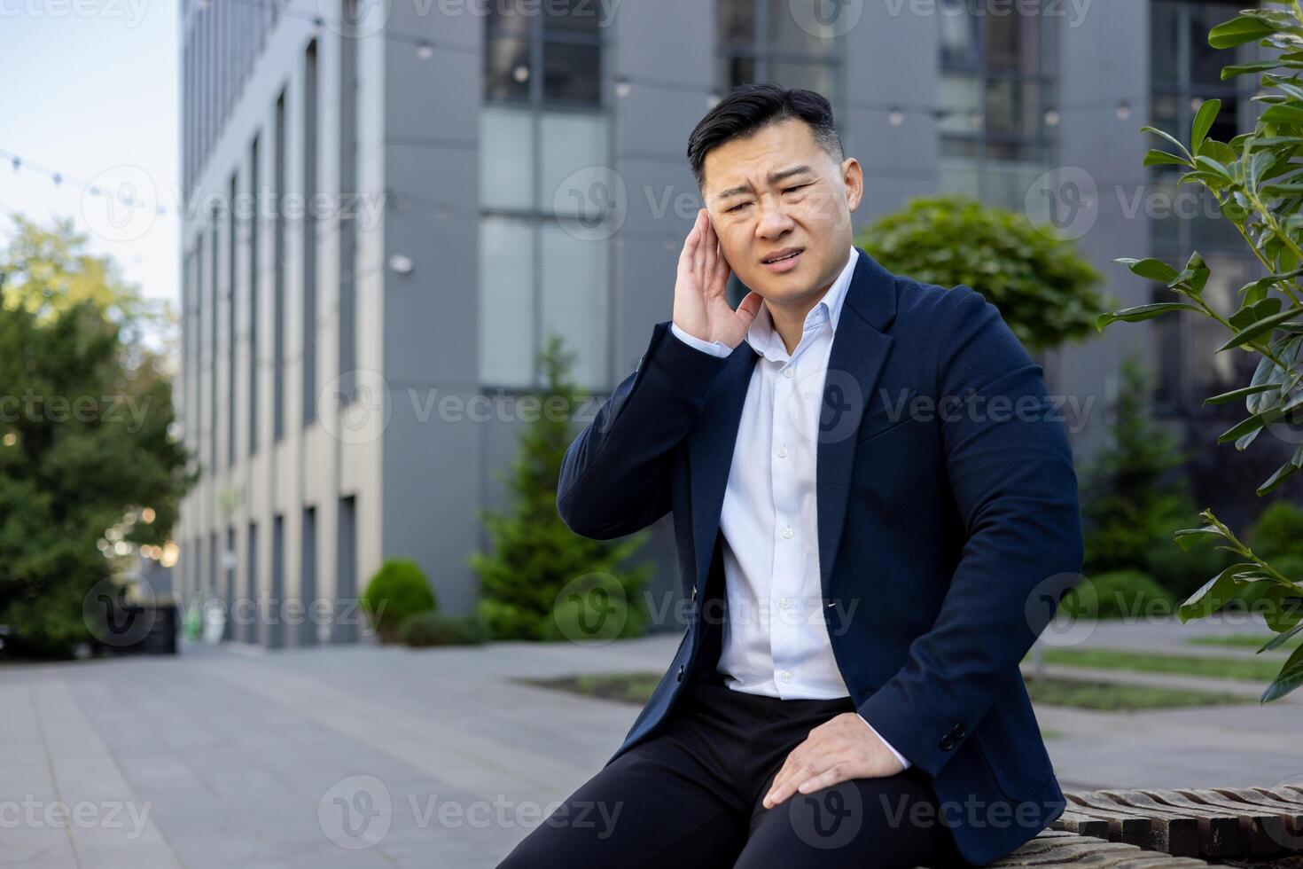 uma jovem ásia homem dentro uma o negócio terno é sentado em uma Banco em a rua perto a escritório Centro, segurando dele mão sobre dele orelha, fazendo careta dentro dor. foto
