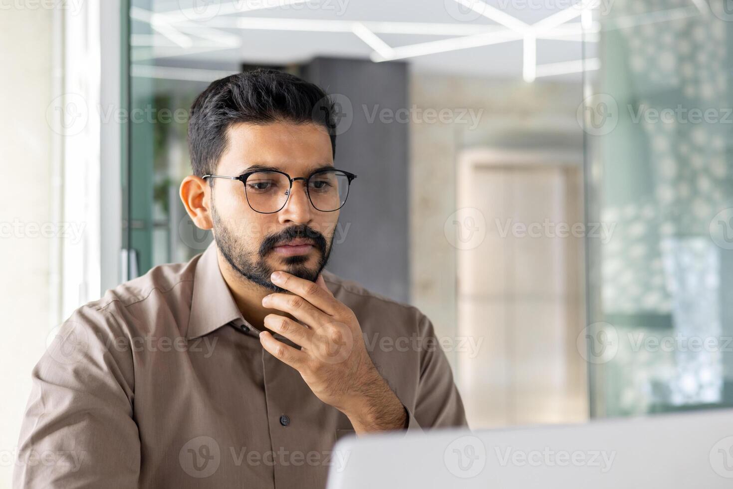 jovem indiano homem de negocios dentro uma sério pensando pose, trabalhando em dele computador portátil às a escritório, mostrando dedicação e foco. foto