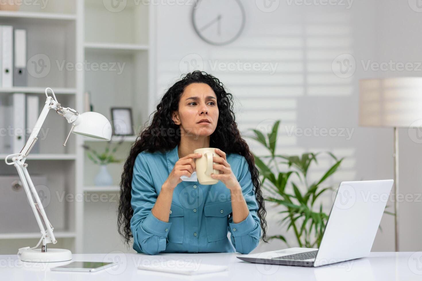 uma mulher dentro uma azul camisa goza uma café pausa às dela limpar, branco escritório espaço com uma computador portátil e interior plantas. foto