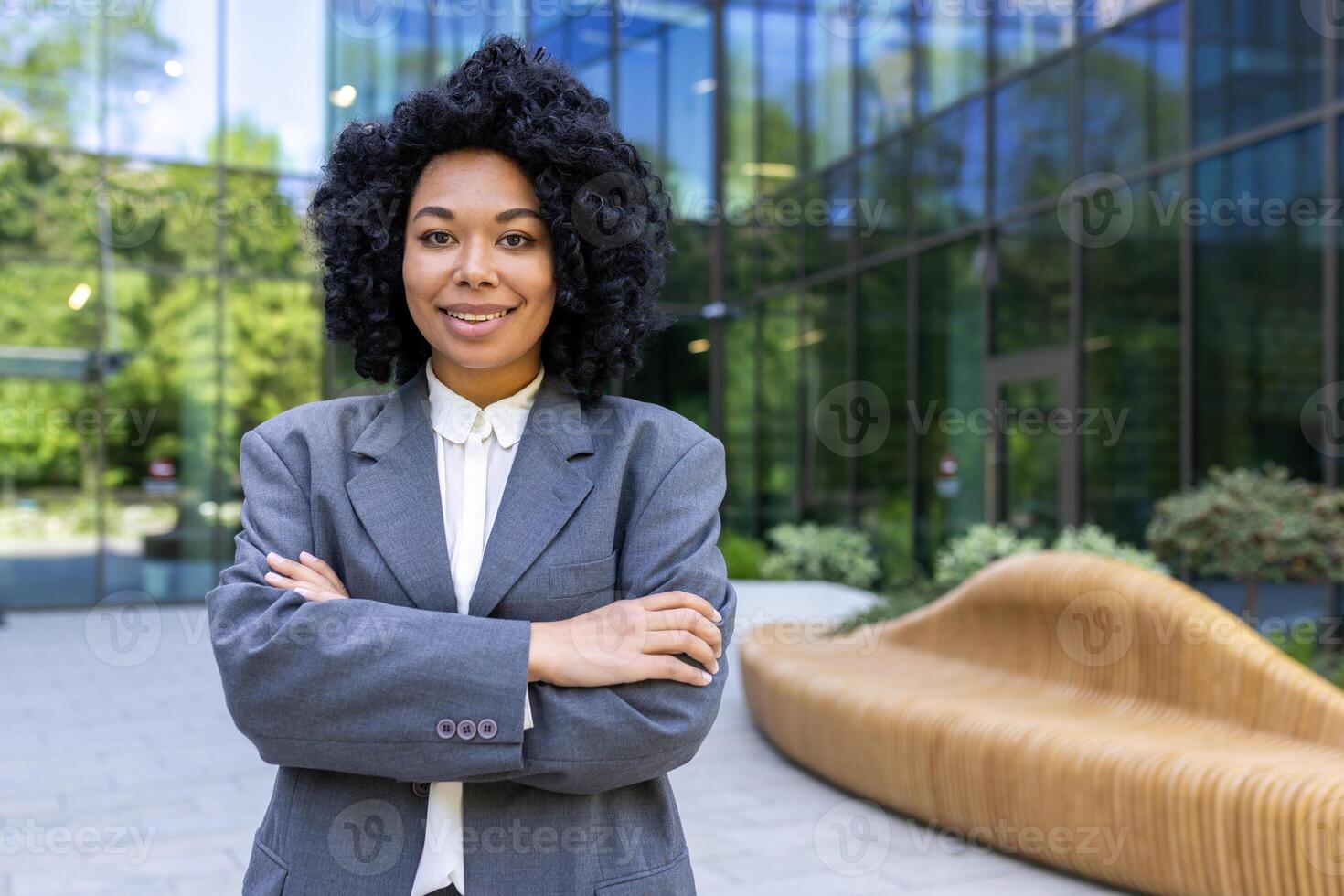 confiante adulto empreendedor dentro roupa formal em pé com guardada mãos em cintura em fundo do moderno prédio. africano americano mulher pronto para Novo o negócio dia e incorporando Liderança e sucesso. foto