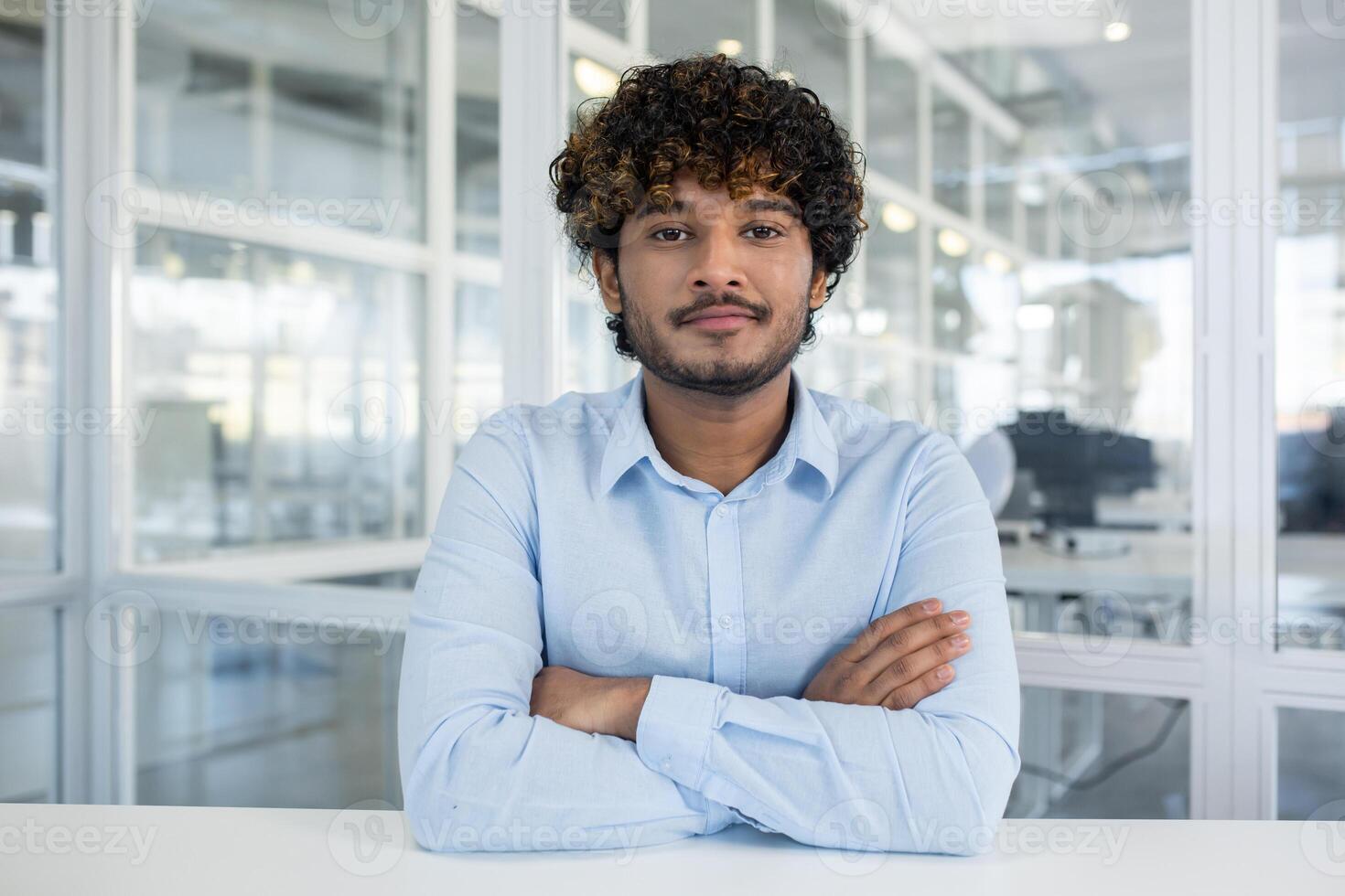 retrato do uma confiante jovem homem com encaracolado cabelo, vestido dentro uma luz azul camisa, sentado dentro uma bem iluminado escritório ambiente. braços cruzado, ele exala profissionalismo e acessibilidade. foto