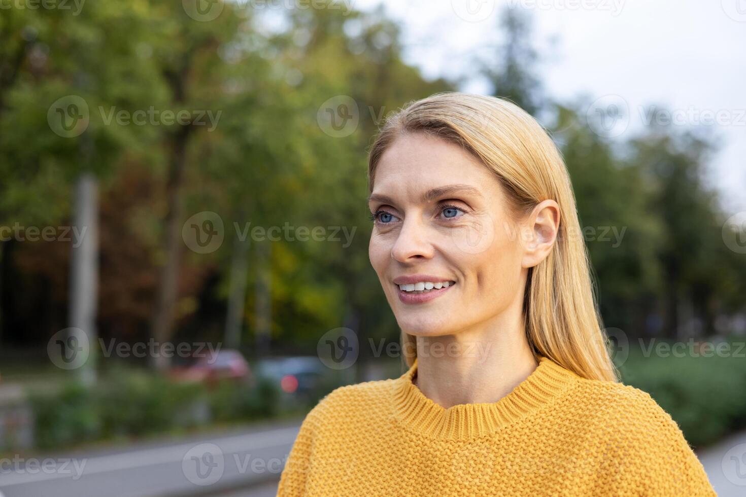 uma alegre meia idade mulher dentro uma brilhante amarelo suéter anda em alegremente dentro a urbano parque contexto, cercado de outono cores. foto