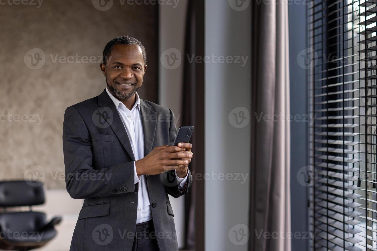 confiante africano homem de negocios dentro uma terno acionado com uma Smartphone enquanto em pé perto a escritório janela. foto