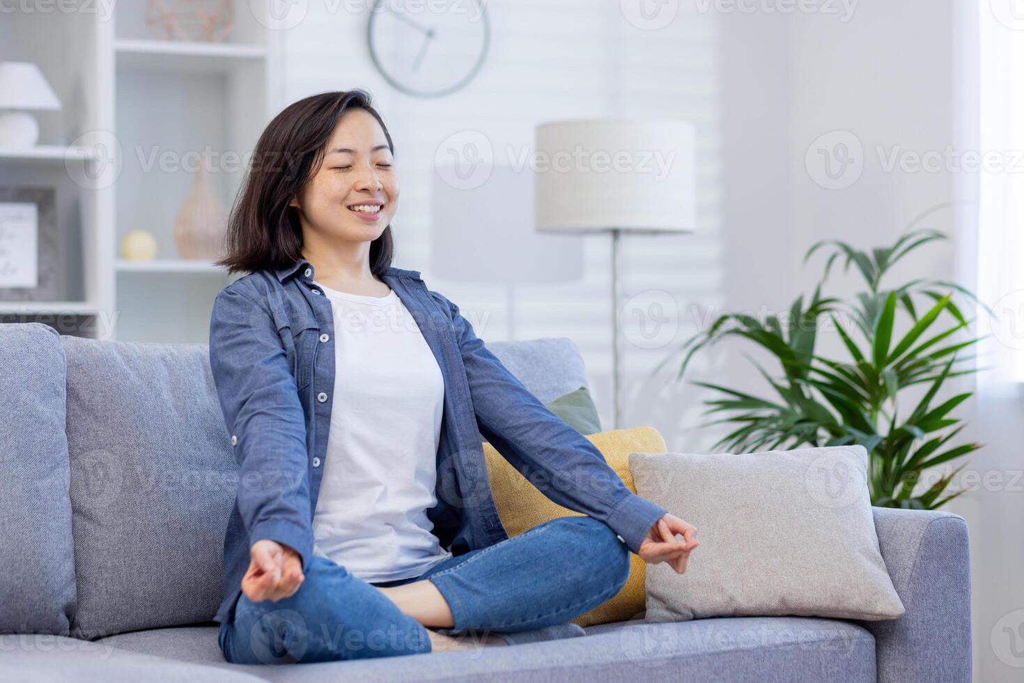 jovem sorridente ásia mulher meditando sentado em sofá dentro vivo sala, mulher sorridente alegremente com olhos fechado, sonhando e pensando sobre futuro planos, visualizando conquista. foto