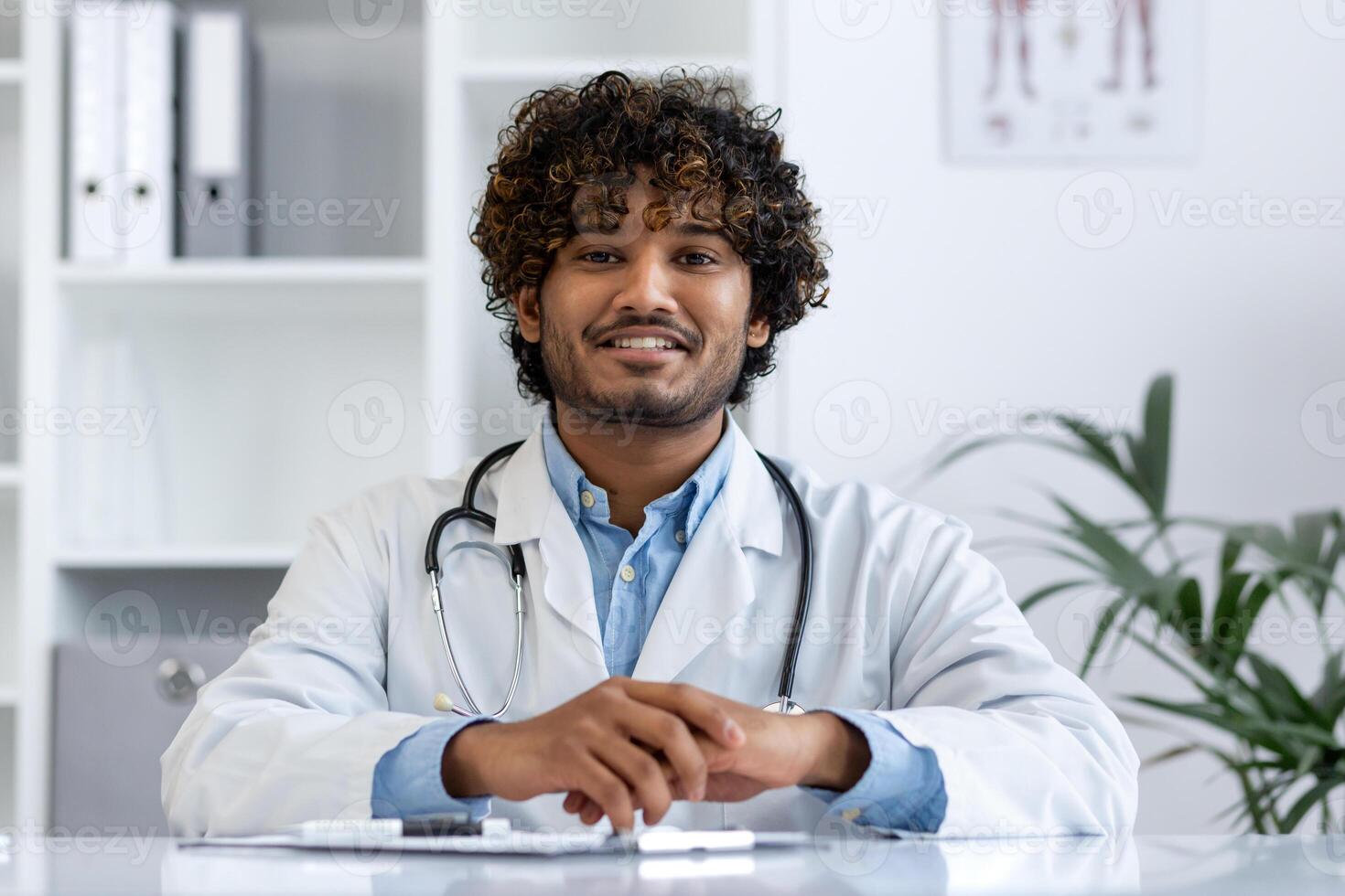 fechar-se retrato do médico dentro clínica às ambiente de trabalho, homem dentro branco médico casaco sorridente olhando às Câmera, satisfeito indiano homem com encaracolado cabelo. foto