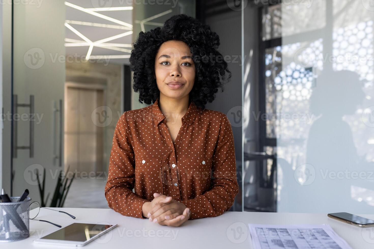 uma confiante jovem africano americano mulher dentro uma à moda polca ponto camisa poses às dela área de trabalho com uma tábua e projeto ,em uma bem iluminado moderno escritório contexto. foto