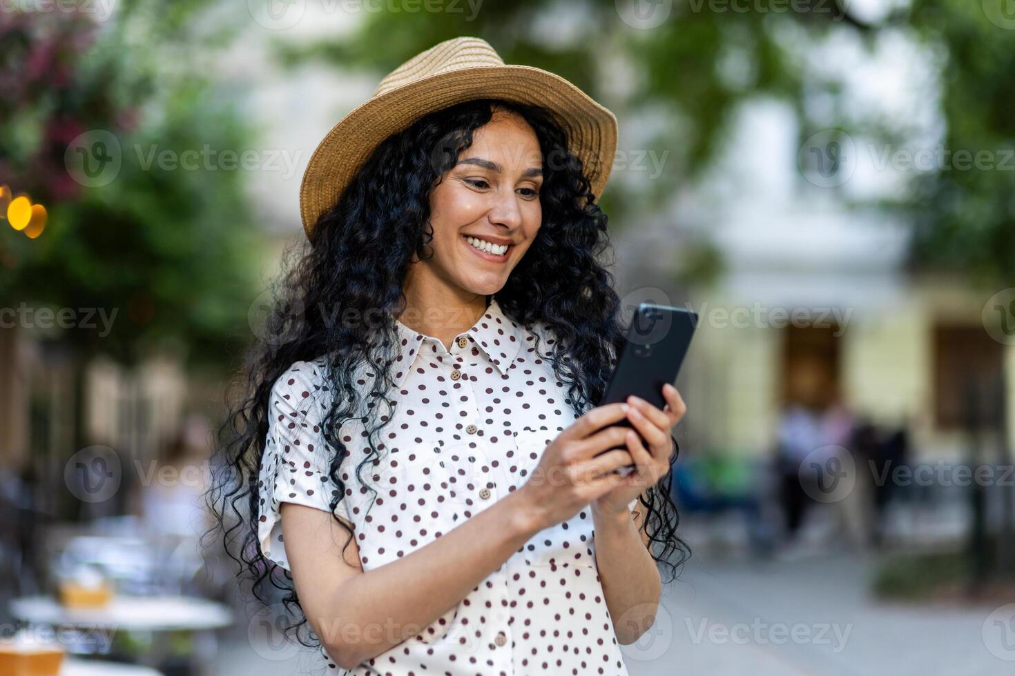 uma lindo jovem mulher anda em através a tarde cidade dentro uma chapéu, uma sorridente latim americano mulher detém uma Smartphone dentro dela mãos. uma turista com encaracolado cabelo tipos uma mensagem e navega conectados Páginas em a telefone. foto