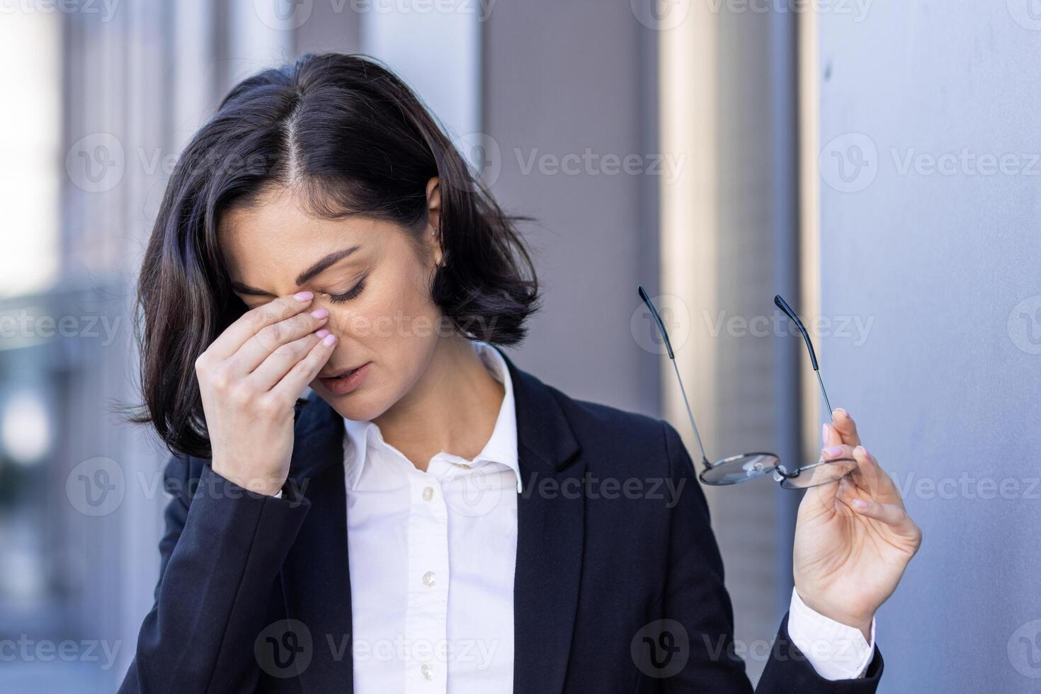 jovem mulher cansado com dor de cabeça, empresária lado de fora escritório construção esfrega olhos, tonto, sobrecarregado trabalhador ao ar livre dentro o negócio terno fechar acima. foto