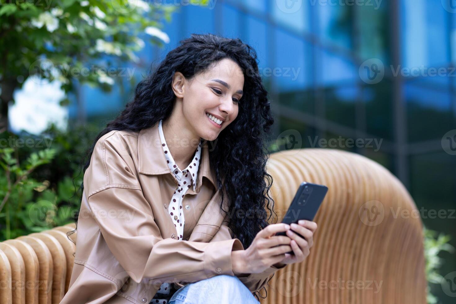alegre lindo mulher sentado em Banco lado de fora escritório prédio, bem sucedido latim americano o negócio mulher usando telefone, lendo notícia conectados e navegando conectados Páginas, segurando Smartphone. foto