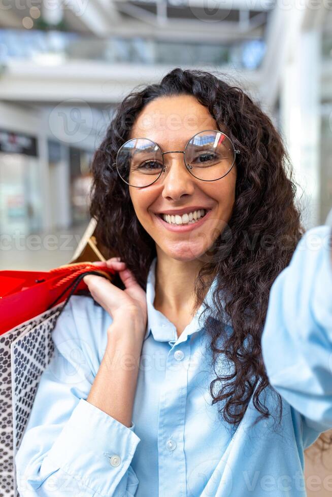 lindo hispânico mulher olhando para dentro Smartphone Câmera falando em ligar e levando selfie foto, compras dentro grande loja segurando colorida presente bolsas dentro mãos sorridente falando com amigos foto