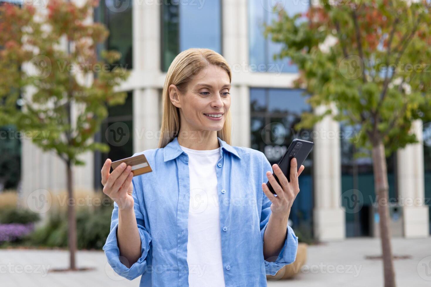 feliz bem sucedido mulher com telefone e banco crédito débito cartão dentro mãos, escolhendo presentes dentro conectados loja, alegremente reserva Serviços e transferindo dinheiro enquanto em pé lado de fora escritório prédio. foto