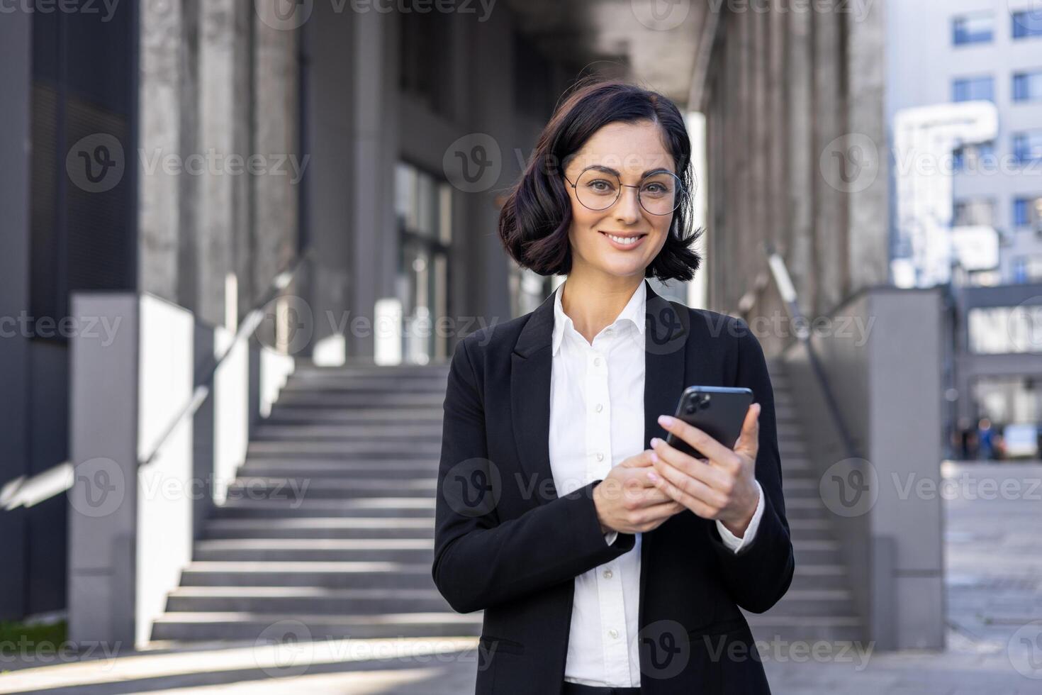 confiante fêmea profissional dentro o negócio vestuário sorridente enquanto usando uma Móvel telefone em uma cidade rua com escadas dentro a fundo. foto