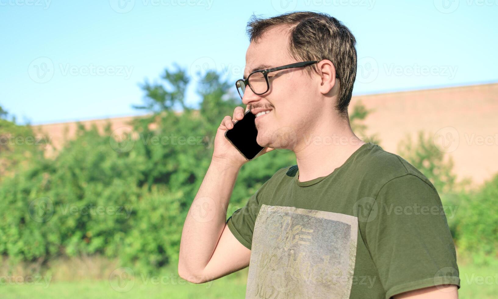 jovem homem com Óculos e verde camiseta falando em célula telefone às a parque foto