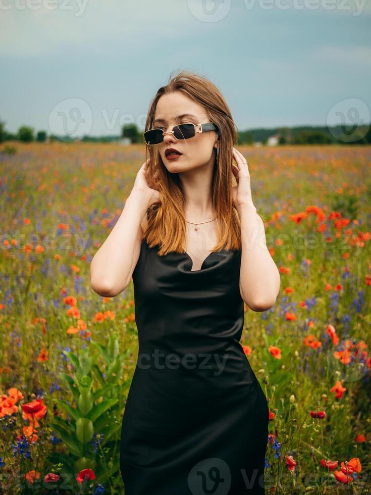 lindo jovem menina dentro uma Preto tarde vestir e oculos de sol posando contra uma papoula campo em uma nublado verão dia. retrato do uma fêmea modelo ao ar livre. chuvoso clima. cinzento nuvens. foto