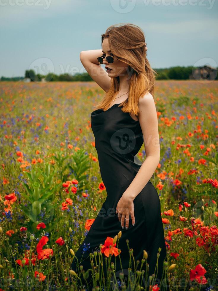 lindo jovem menina dentro uma Preto tarde vestir e oculos de sol posando contra uma papoula campo em uma nublado verão dia. retrato do uma fêmea modelo ao ar livre. chuvoso clima. cinzento nuvens. foto
