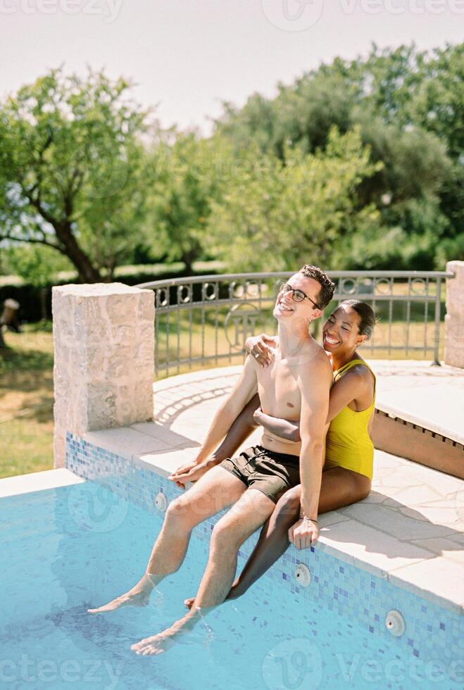 sorridente mulher abraços homem a partir de atrás enquanto sentado com ele em a Beira do uma natação piscina foto