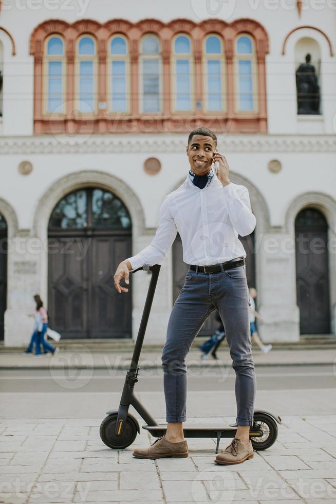 jovem afro-americano usando telefone celular em pé com uma scooter elétrica em uma rua foto