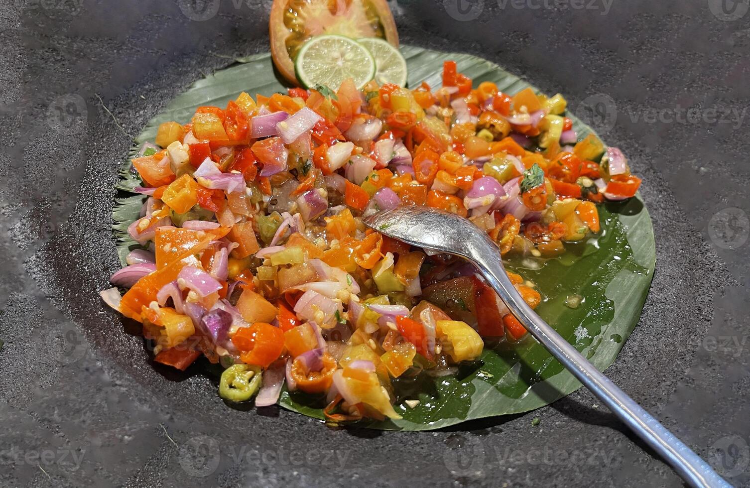 picante condimento do indonésio sambal dabu dabu fez do Pimenta, tomate e chalota com limão manjericão dentro uma cerâmico tigela, de madeira colher em cinzento fundo foto