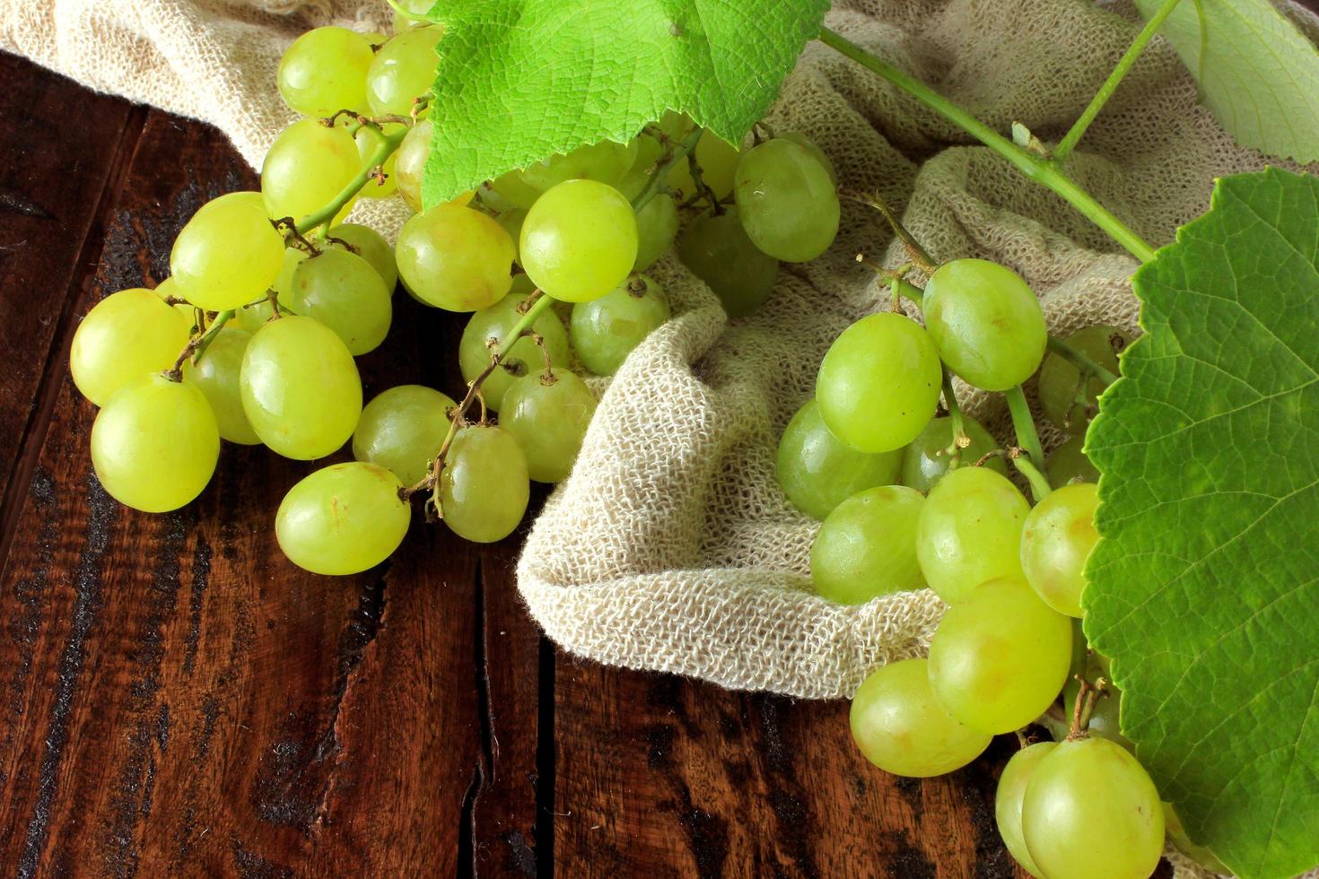 cachos de uvas verdes frescas, com galhos e folhas, tecido rústico, em mesa de madeira rústica foto