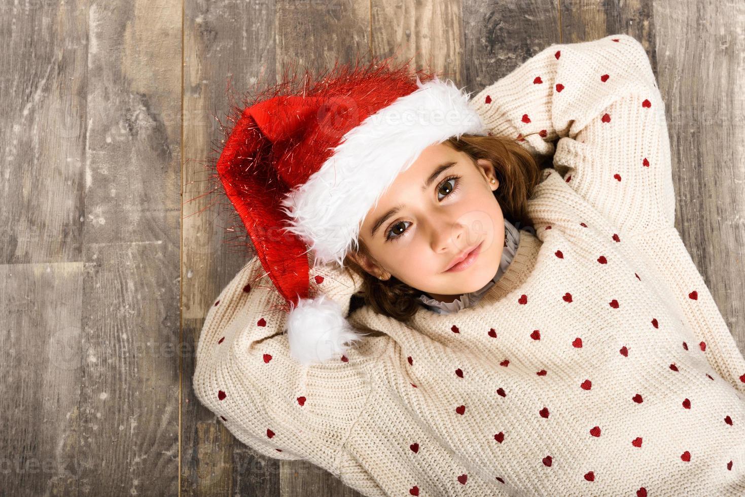 adorável menina com chapéu de Papai Noel deitada no chão de madeira foto