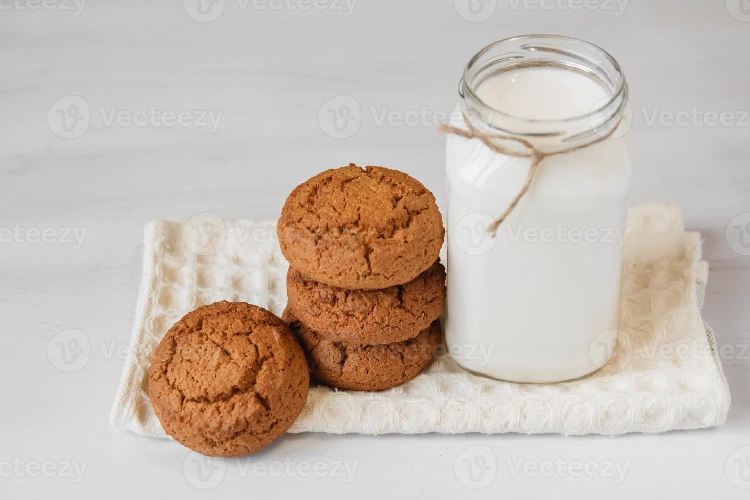 leite em uma jarra de vidro e biscoitos de aveia perto do guardanapo na mesa branca foto