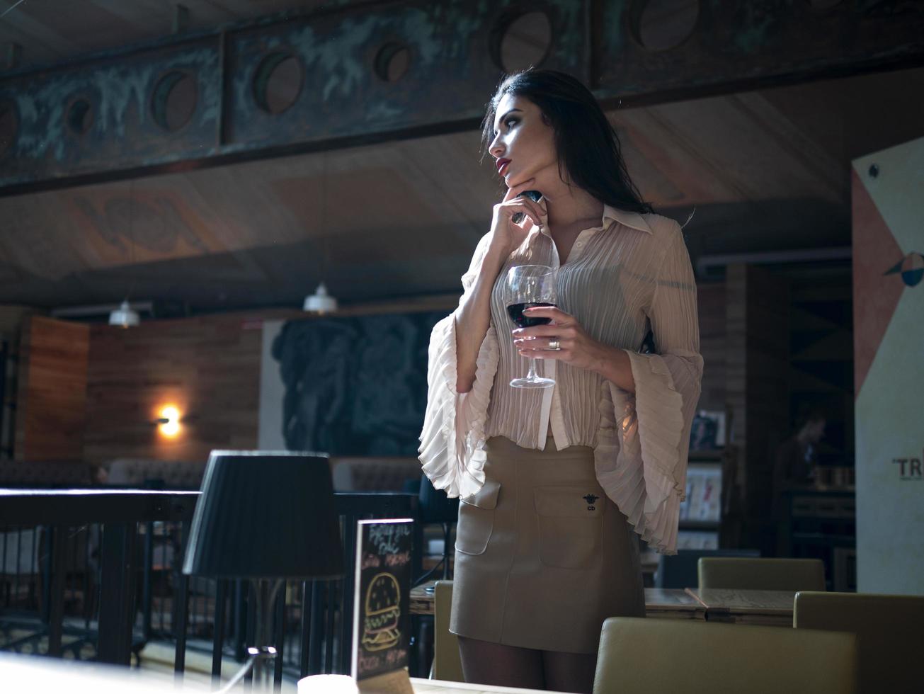 modelo de garota linda posando de pé com uma taça de vinho tinto na mão em um restaurante foto