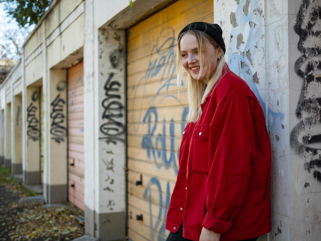 jovem com chapéu de malha e jaqueta vermelha sorrindo em pé perto da parede com grafite foto