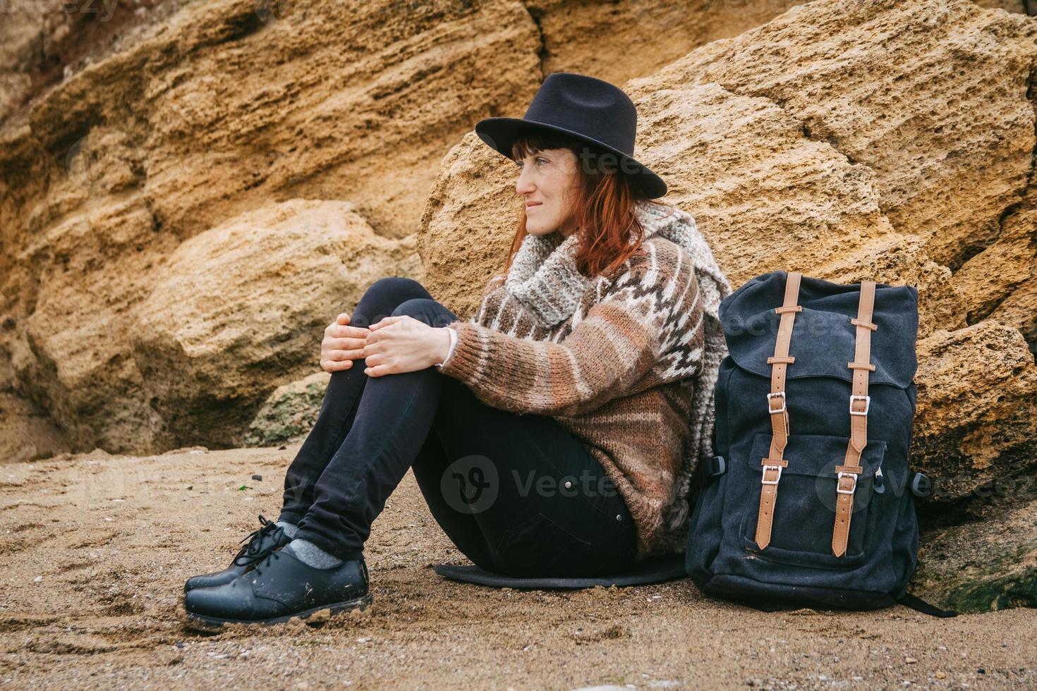 mulher com um chapéu e um lenço com uma mochila sentada na costa sobre um fundo de pedras foto