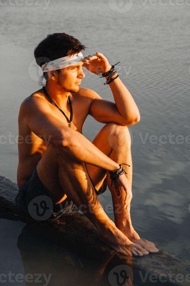 homem com músculos perfeitos sentado na ponte de madeira e olhando para o reflexo da água ao pôr do sol foto