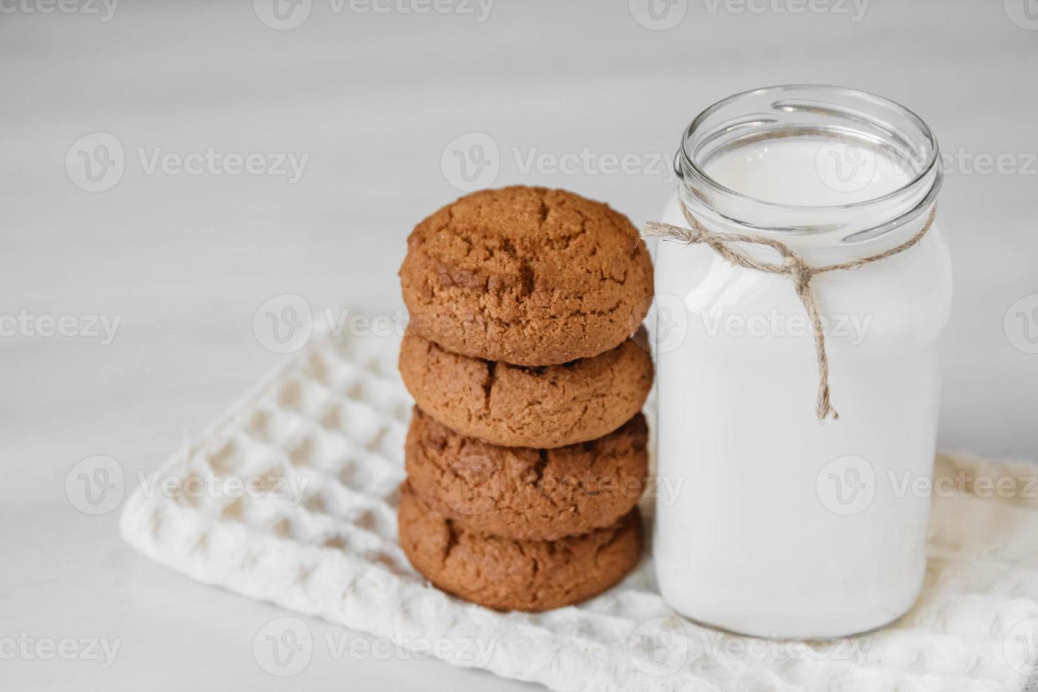 leite em uma jarra de vidro e biscoitos de aveia perto do guardanapo na mesa branca foto