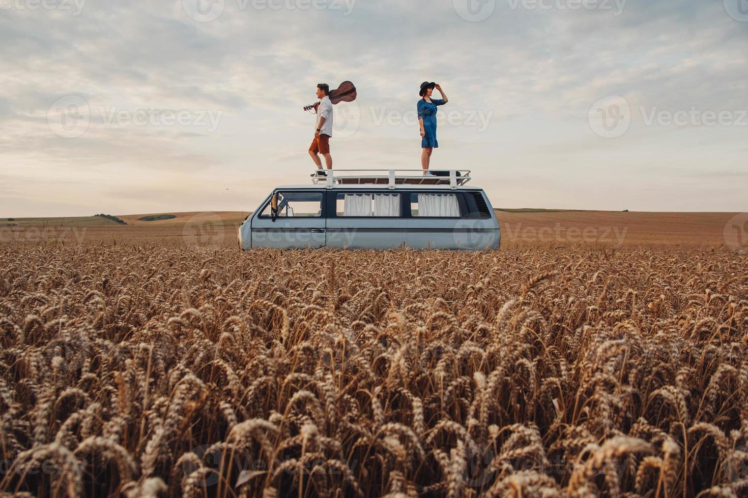 casal homem com uma guitarra e uma mulher com um chapéu estão de pé no telhado de um carro em um campo de trigo foto