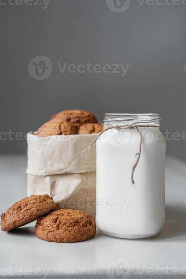leite em uma jarra de vidro e biscoitos de aveia em um saco de papel sobre um fundo de mesa branco foto