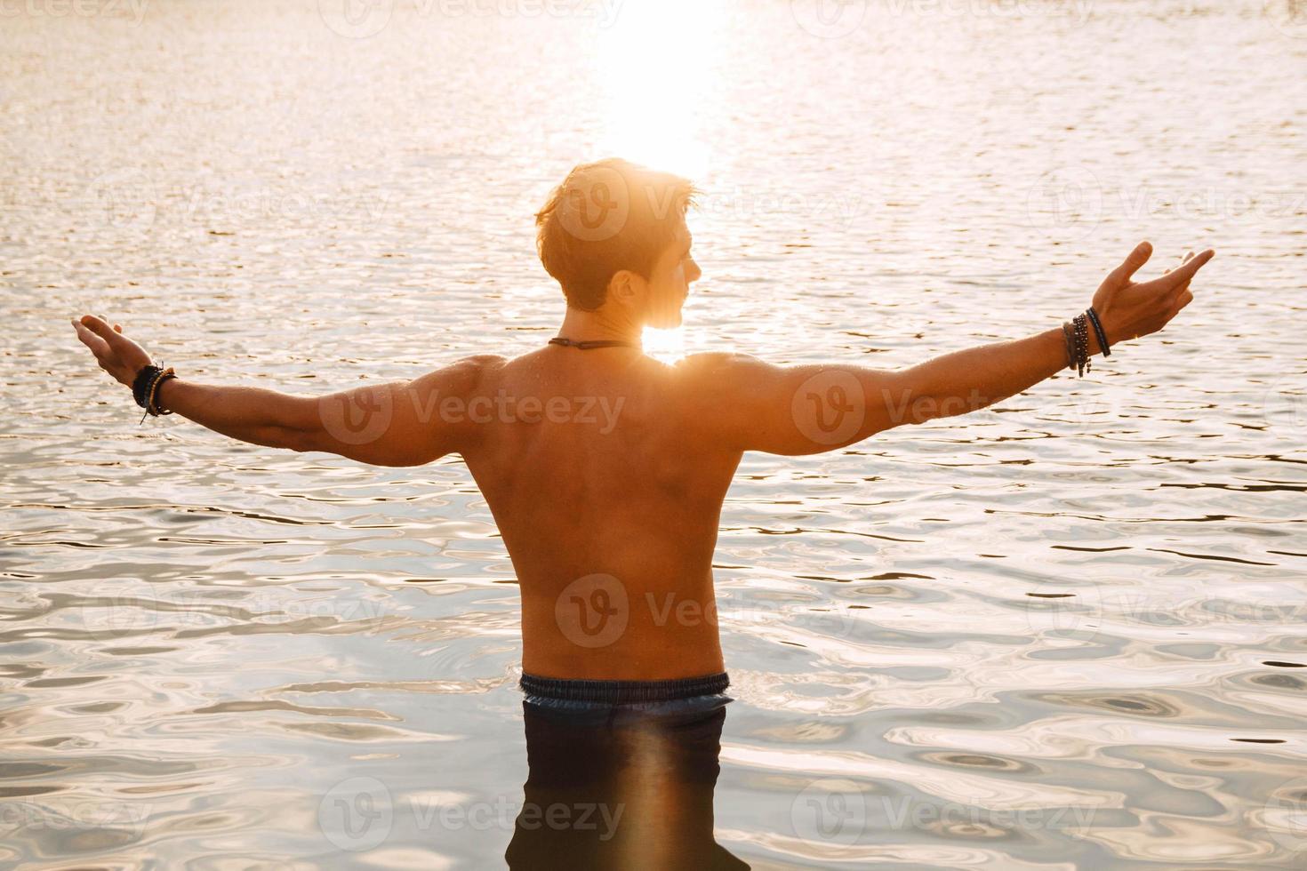 homem com músculos perfeitos na água ao pôr do sol, jogando as mãos para o lado foto